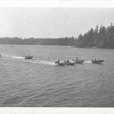 Seven boats participating in a boat race on American Lake, the tree-lined shores viisble behind