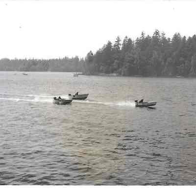 Three boats participating in a boat race, a fourth just visible on the left, tree-lined shores behind