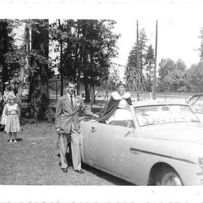 Queen Rita Gardner sitting on car in preparation for 4th of July parade with man and child standing outside car