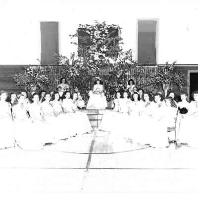 Four rows of girls in white sitting in a v-formation in front of three other girls on a podium