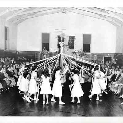 Girls in white dance at the end of maypole tethers inside in front of a seated crowd