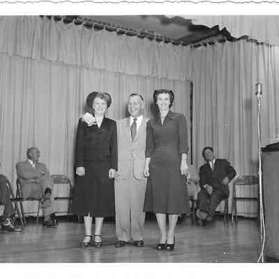 A. G. Hudtloff with his arms around Mrs Alton and Mrs Jeffries while standing on a stage with people and curtains behind and a podium and standing microphone in front