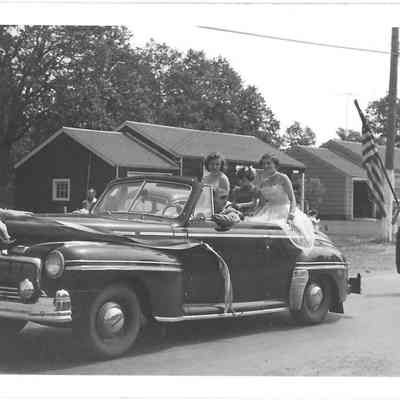 Two Tillicum pageant girls, Nancy Ward and Rita Gardner, alongside flower girl Pamela Ried riding in black convertible followed by girl with flag