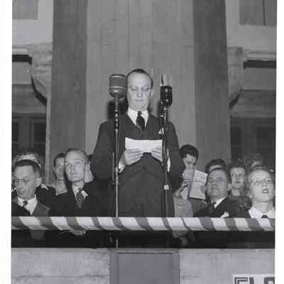 A. G. Hudtloff reading reading piece of paper in front of two microphones at Clover Park, small crowd behind him
