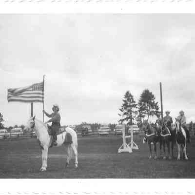 Four young women on horse back, one apart from the rest raising an American flag