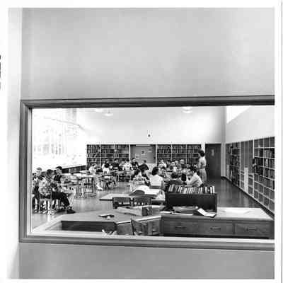 Room full of students studying at desk and bookshelves as seen through a window in the hallway outside at Hudtloff Junior High