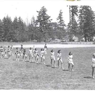 Seventeen or so young people with batons participating in Grand March through field, baseball field, cars, trees, and buildings in distance