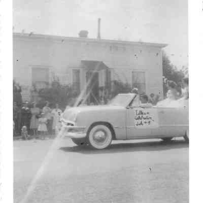 Pale convertible with two pageant girls in back and a Tillicum Celebration sign on side pass by pale building and crowd