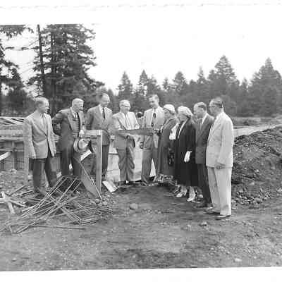 Nine people standing in dirt field with trees in background as a man in the middle appears to pass a saw to the man on his right