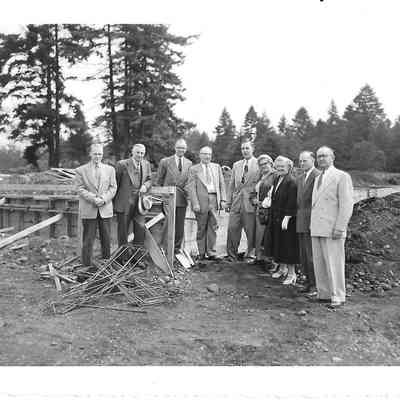 Nine people standing in a dirt field with trees in background with one man leaning on small wooden structure