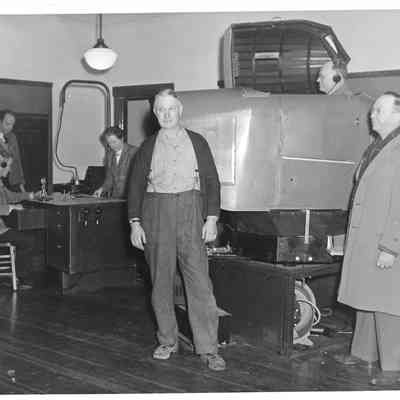 A. G. Hudtloff and another man posing in front of a model cockpit with a man inside while four people stand around a desk in the background