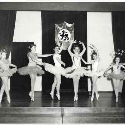 Six girls dancing ballet on a stage in front of a crested banner: 2010.013.004.c