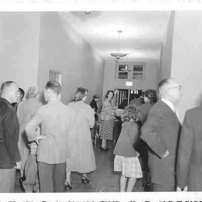Backs of several adults and children standing in a crisp looking hallway while one woman in the middle looks directly at camera
