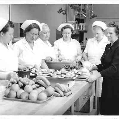 Six women making food on butcherblock table, large pile of fruit in front