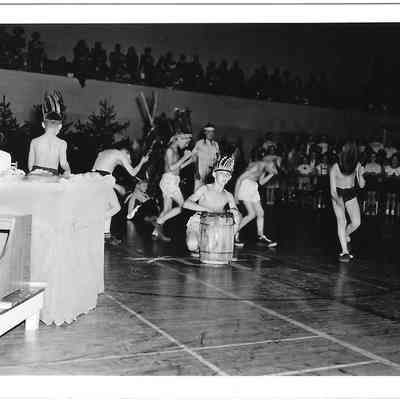 Children wearing headdresses, dancing, and playing drums in middle of gym assembly
