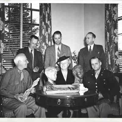 A. G. Hudtloff and Pearl A. Wanamaker in room with four others behind table with two busts while others hold wood carved figures