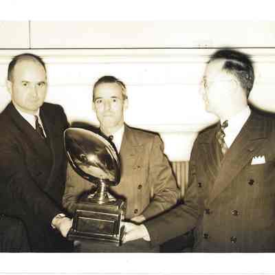 Bill Hewett, Henry Jorkins, and A. G. Hudtloff holding a football trophy while Hugh Johnson sits in front of them