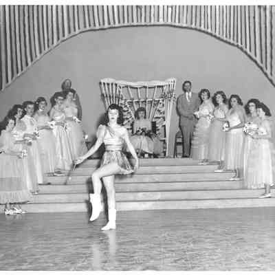 Baton twirler dances before pagent queen on her throne and her court of princesses