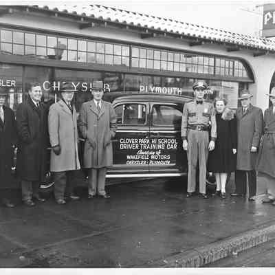 A. G. Hudtloff and seven others standing in front of a car with Clover Park Hi School Driver Trianing Car printed on it and the Plymouth Chrysler building