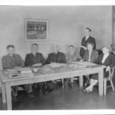A. G. Hudtloff stands looking down at four men and two women all sitting at a desk covered in papers