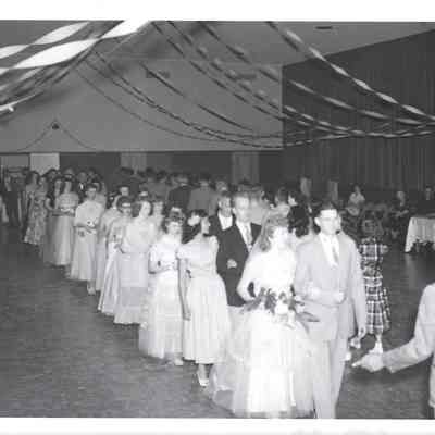 Line of young women arm-in-arm with adult men approaching man with arm outstretched beneath streamers