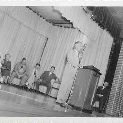 Man in light shaded suit talking forward at podium near standing mic while several people sit in front of curtains behind him