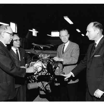 Four men including Wallace B. Hagen inspecting an engine with a car in the the background