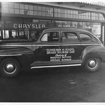 The left side of a car with Clover Park Hi School Driver Trianing Car printed on it parked in front of the Plymouth Chrysler building