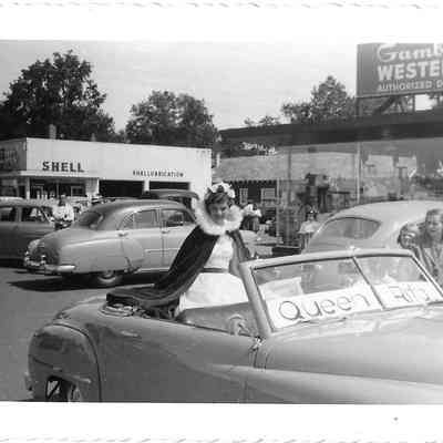 Rita Gardner on back of convertible in front of a Shell station and Gambles Western dealership
