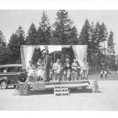 Seven people including pageant queen sat on small outdoor stage with one additional man standing at mic, possible on field