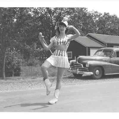 Nancy Jo Dross in Majorette outfit twirling baton in front of parked car and house