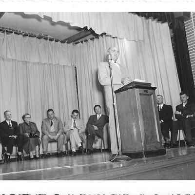 Man in light shaded suit talking to his right while at podium near standing mic while several people sit in front of curtains behind him 1950
