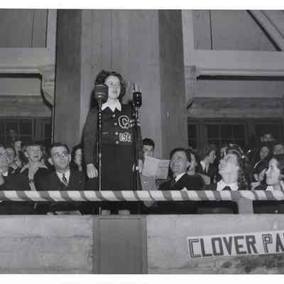 Unidentified girl in Clover Park letter sweater in front of two microphones at Clover Park, small crowd behind him