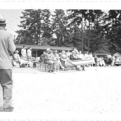 Crowd sitting outdoors in chairs, front line holding a partially visible flag, man in foreground on left