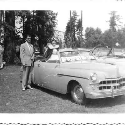 Queen Rita Gardner sitting on car in preparation for 4th of July parade with man standing to her right