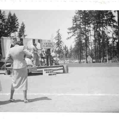 Man on small outdoor stage holding up sing at the dedication of the H.M. Bona Playfield , two women in foreground on left