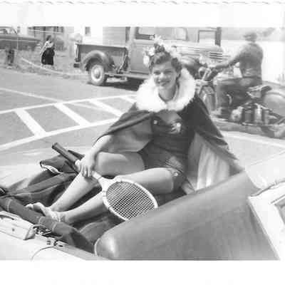 Rita Gardner sitting on back of car, wearing crown, cape, and holding a tennis racquet