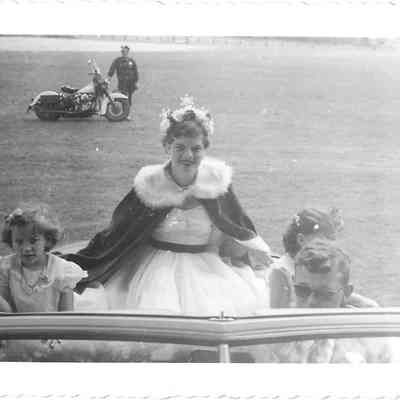 Rita Gardner on the back of a car wearing crown and cape, flanked by two young girls with a man in glasses driving parade car