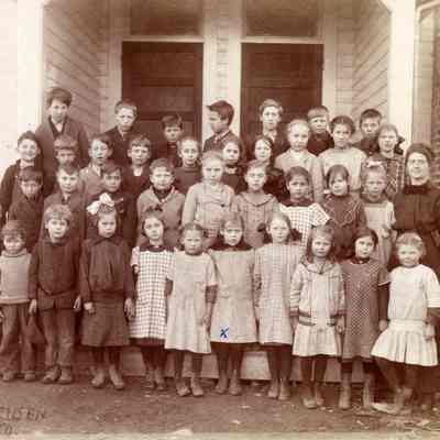 Black and white photograph of a group of schoolchildren and their teacher, gathered in front of a schoolhouse building: 2023_001_I_F_a04 photo by H.G. Sorensen
