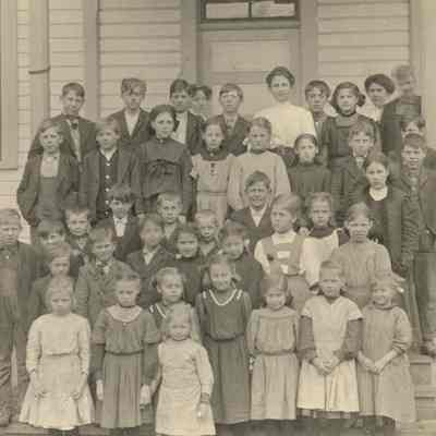 Black and white photograph of a group of schoolchildren and their teacher, gathered on the front steps of a schoolhouse: 2023_001_I_F_a05