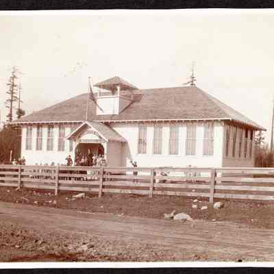 Black and white photograph of the Livingston Bay Schoolhouse from across the road with some schoolchildren in front of the building: 2023.001.I.F.c02