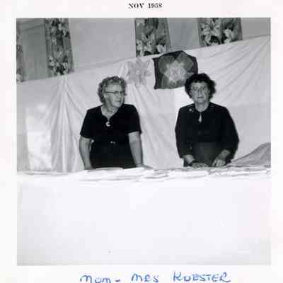 Mom, Mrs Robster, Nov 1958: A black and white photograph of two women standing behind a table with what is possibly lace tablecloths, doilies, or curtains on it. 2023.001.V.011