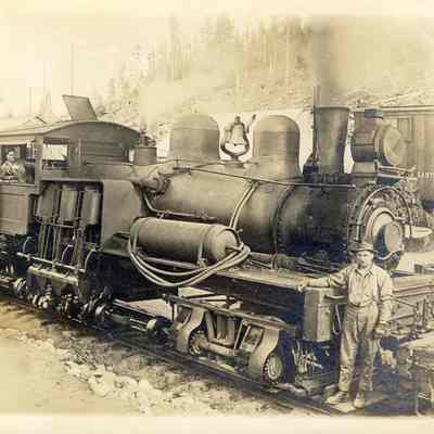 A train engine with three people.: Two figures stand inside a train engine car - and one stands on the front end - which sits on train tracks. There do not appear to be any cars attached. 2023.001.I.B2.a011