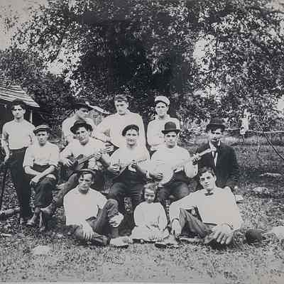A group of male Retaliate family members, with some playing musical instruments, pose on their farm beside a child.