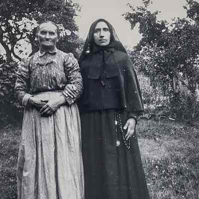Two women in traditional clothing stand together outdoors, one holding a rosary,