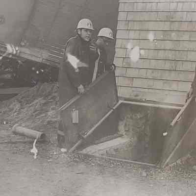 Workers in helmets inspect an open cellar door near a house, amidst a construction site scene.