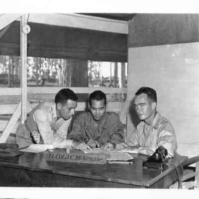 Photo of three men in office. The sign on the desk says Lt. Col. J. C. McKenzie