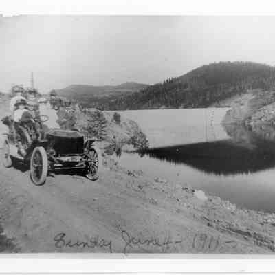 3. Photo of Stanley Steamer with passengers at east end of Barker Reservoir. Violet Norris Forbes identified herself as the high school girl in the front seat. Mrs. Wolcott was standing behind her. Mrs. J.G. Clark was the woman in the white dress in the back seat with Mrs. Ed Norris. She thought her brother, Prentiss Norris, was standing beside Mrs. Wolcott.: 5"x7" black and white photograph