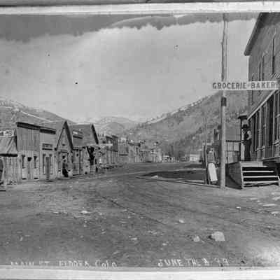 5. Looking West, Main St., Eldora, Colo., June the 8 - 99: Main Street in Eldora in 1899 shows (left side) the Mogul House - Meals, Livery Feed & Sale Stable. On the right side is the Grocerie and Bakery and the Post Office. The photo also shows several men, one woman with a white apron, and a dog.  Back of photo: "Mogul House- Mrs. Jamieson, children Danny, Molly, Frances. Grocerie & Bakery and Hotel Wardell". 5"x7" print located in 2024.10.60 page 7