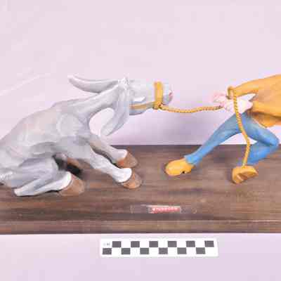 "Stubborn" - Painted wood carving of mule sitting on haunches being pulled by a man